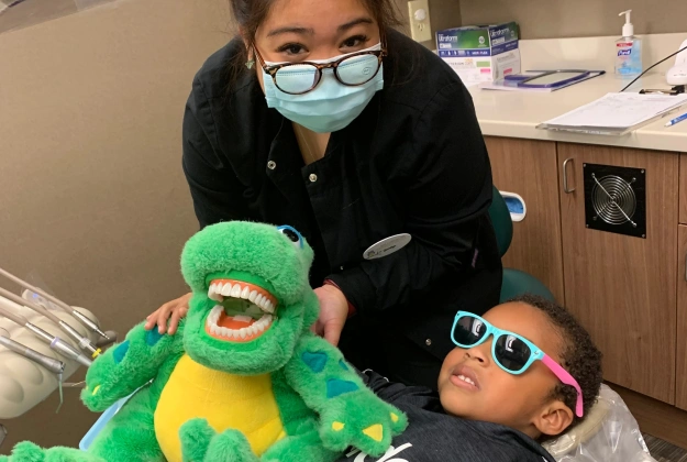 Pediatric dentist wearing mask holds green puppet while child wears sunglasses in dental office.