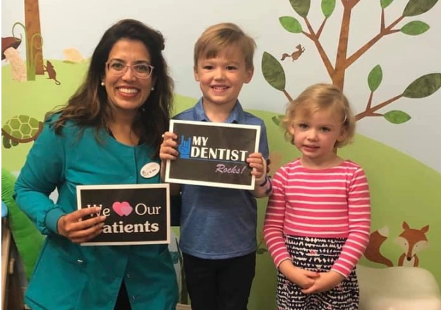 Female dentist with two young children holding dental educational materials in office setting.
