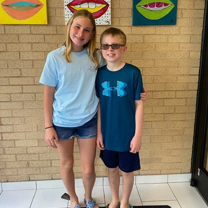Two children smiling at camera in dental office with colorful artwork on brick wall behind them.