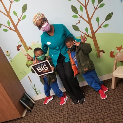 Pediatric dentist with child holding "Big" sign in front of tree mural in dental office.