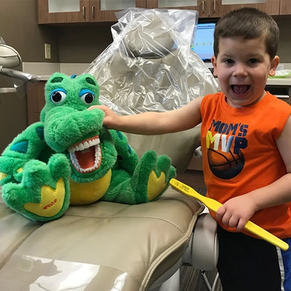 Young child in orange shirt smiling while holding green plush alligator toy in dental office.
