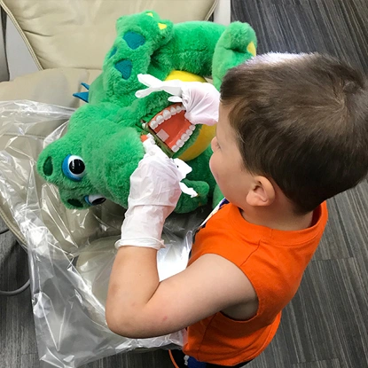 Child in orange shirt holding green plush dinosaur toy in dental office setting.