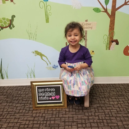 Young child in purple shirt smiling next to first day of school sign in pediatric dental office.