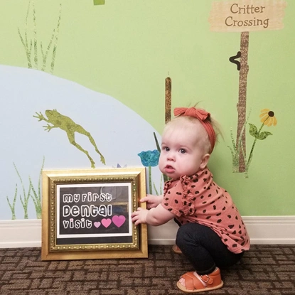 Toddler in pink outfit sitting next to chalkboard sign and toy frog in dental office setting.