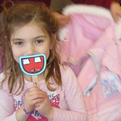 Young girl holding decorative smile prop in front of face, wearing pink shirt in dental office setting.