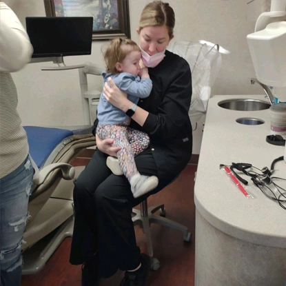 Pediatric dentist holding smiling toddler in dental office treatment room.