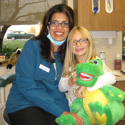 Pediatric dentist in blue scrubs holding green plush toy with young girl wearing glasses in dental office.