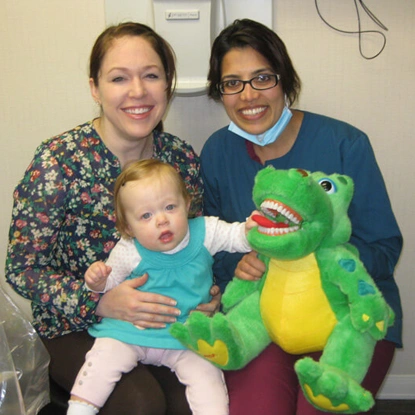 Two adults and a toddler holding a green dinosaur toy in a dental office setting.