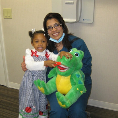 Woman and child holding green plush toy in dental office setting.