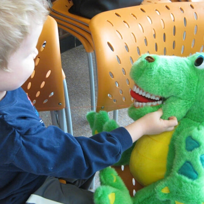 Young child in blue shirt touches green frog puppet in dental office waiting area.