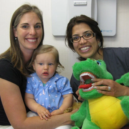 Two women and a toddler hold a green dinosaur toy in a pediatric dental office.