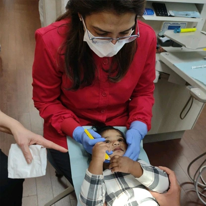 Pediatric dentist in red shirt and blue gloves examining young child's teeth in dental office.