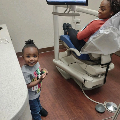 Young child smiling at dental office with dentist and assistant in background near chair.