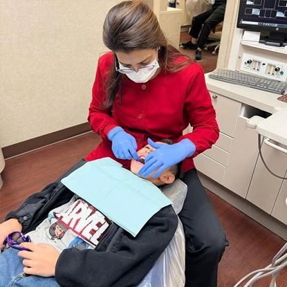 Pediatric dentist in red shirt and mask examines young patient in dental office setting.