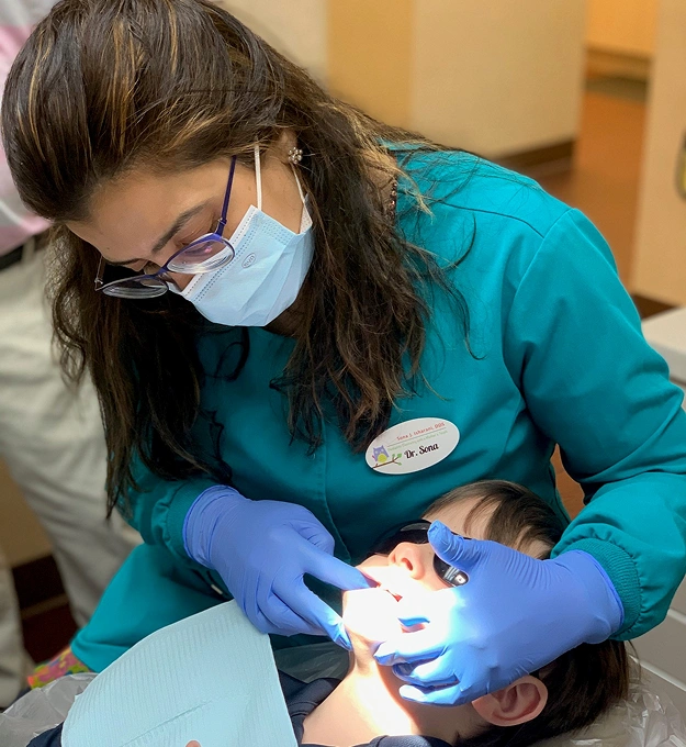 Pediatric dentist in blue scrubs examining young child's teeth in dental office.