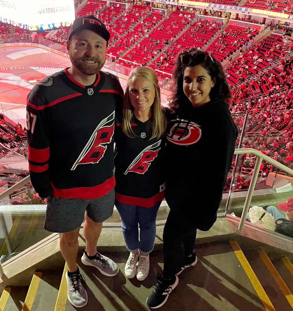 Three people wearing red and black sports apparel stand in a stadium luxury box during a game.