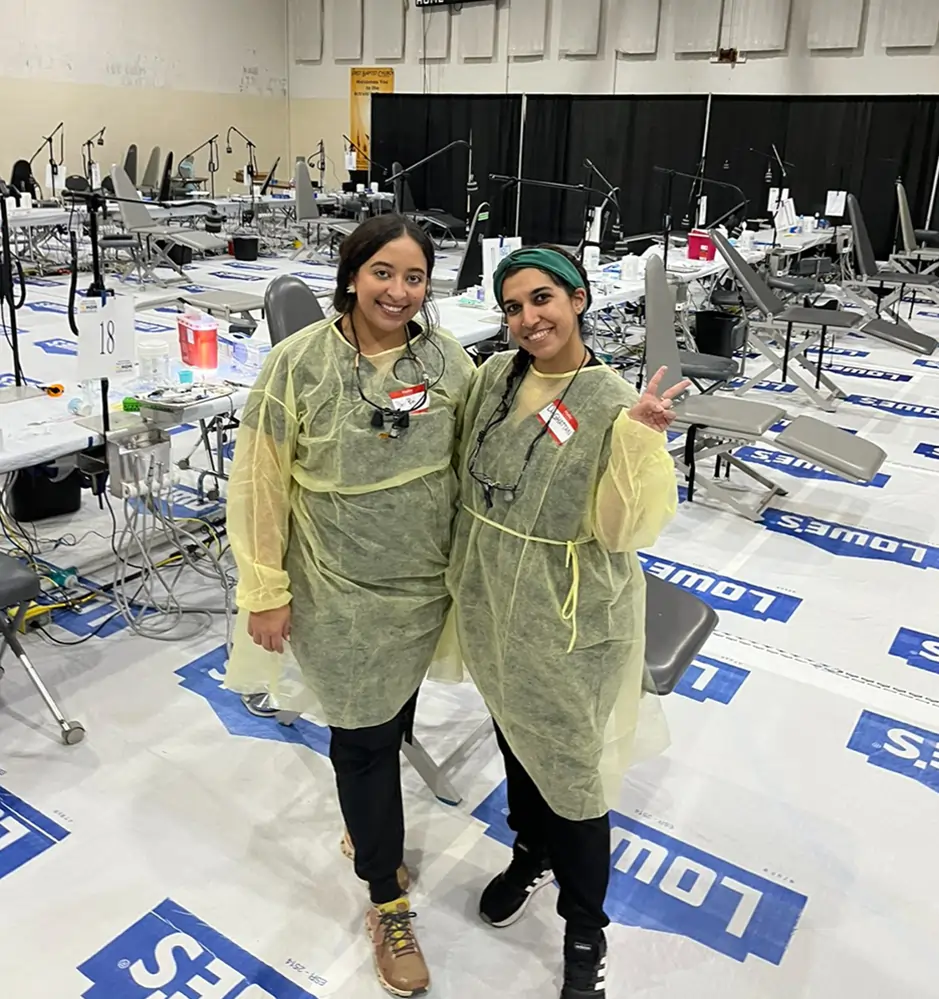 Two women in yellow protective gowns pose together in a large event venue with blue flooring.