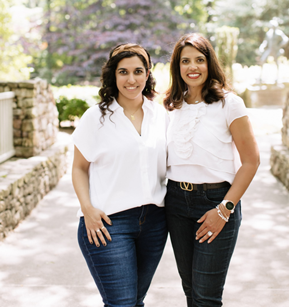 Two women in white shirts and dark pants standing together outdoors on a stone pathway.