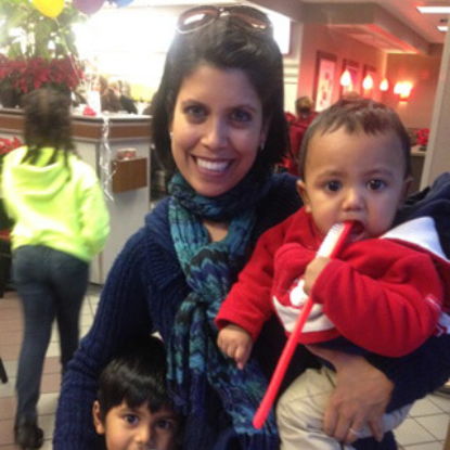 Woman holding toddler in red jacket at indoor event or clinic setting with child nearby.