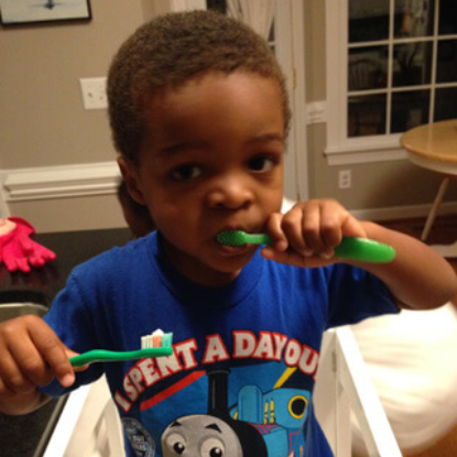 Young child in blue shirt brushing teeth with green toothbrush indoors.