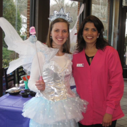 Two women pose together indoors; one wears a white angel costume with wings and tiara, the other wears pink.