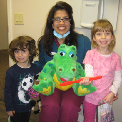 Three children posing with dental professional holding green dinosaur puppet in dental office.