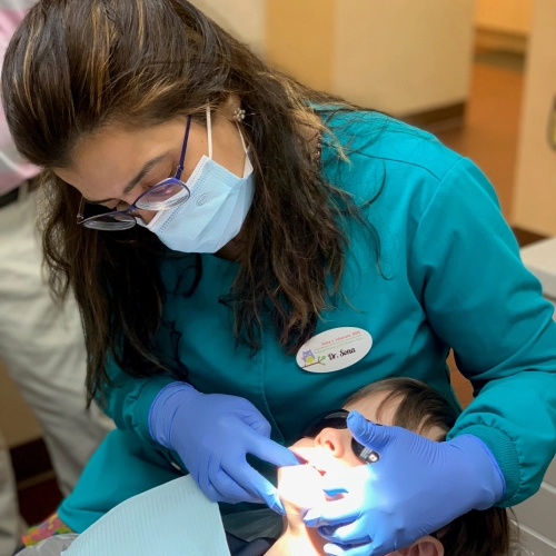 Pediatric dentist in teal scrubs and mask examines young child's teeth in clinical setting.