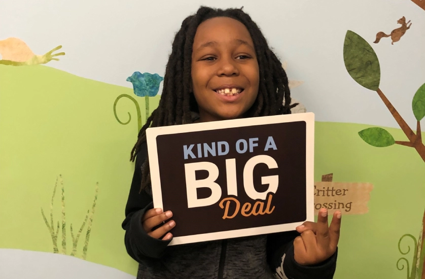Young girl smiling while holding "Kind of a Big Deal" sign in dental office setting.