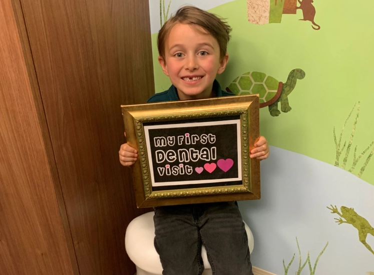 Child holding framed award in dental office with green wall and dinosaur decorations.